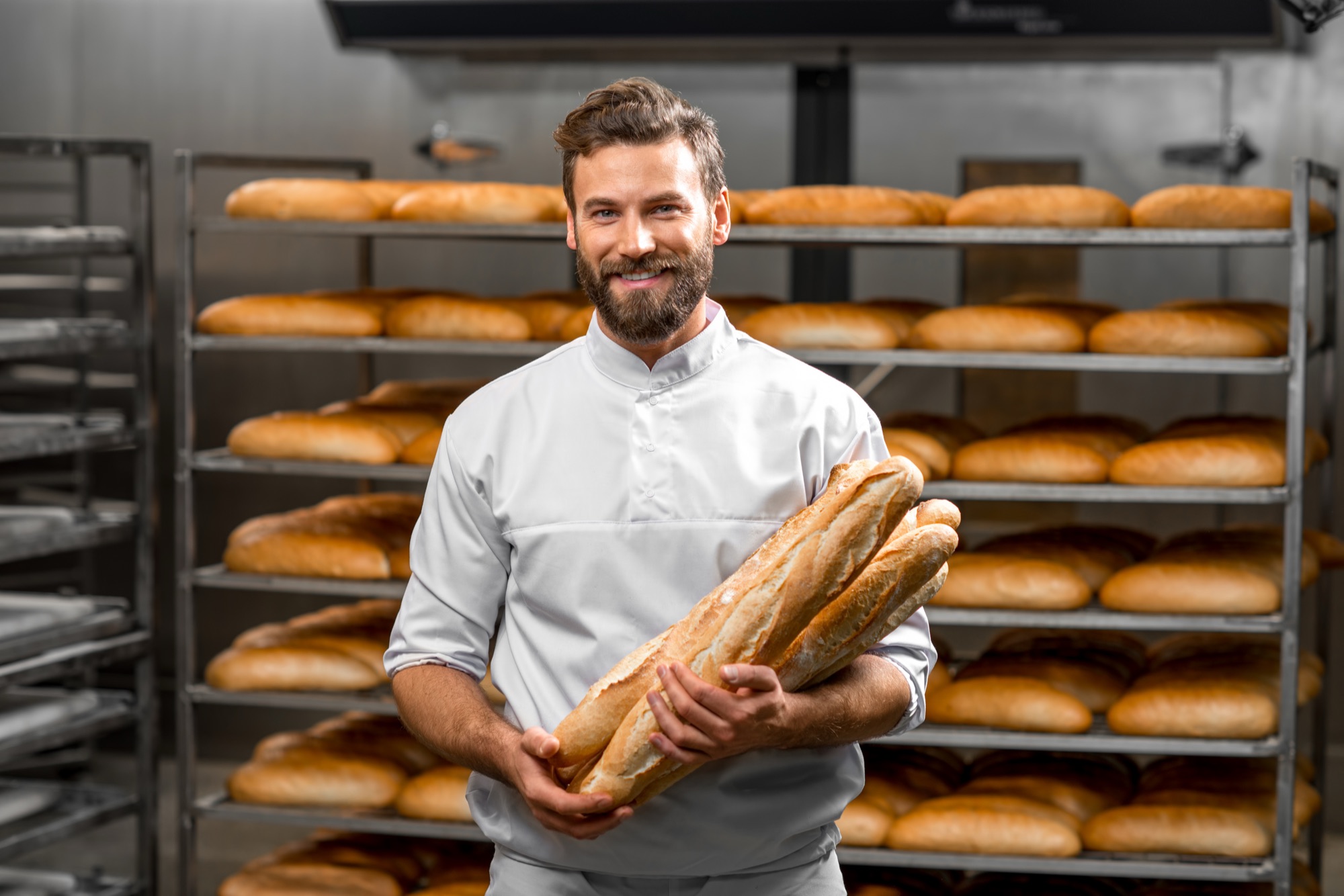 Bäcker hält frische Baguettes vor dem Gestell mit Broten